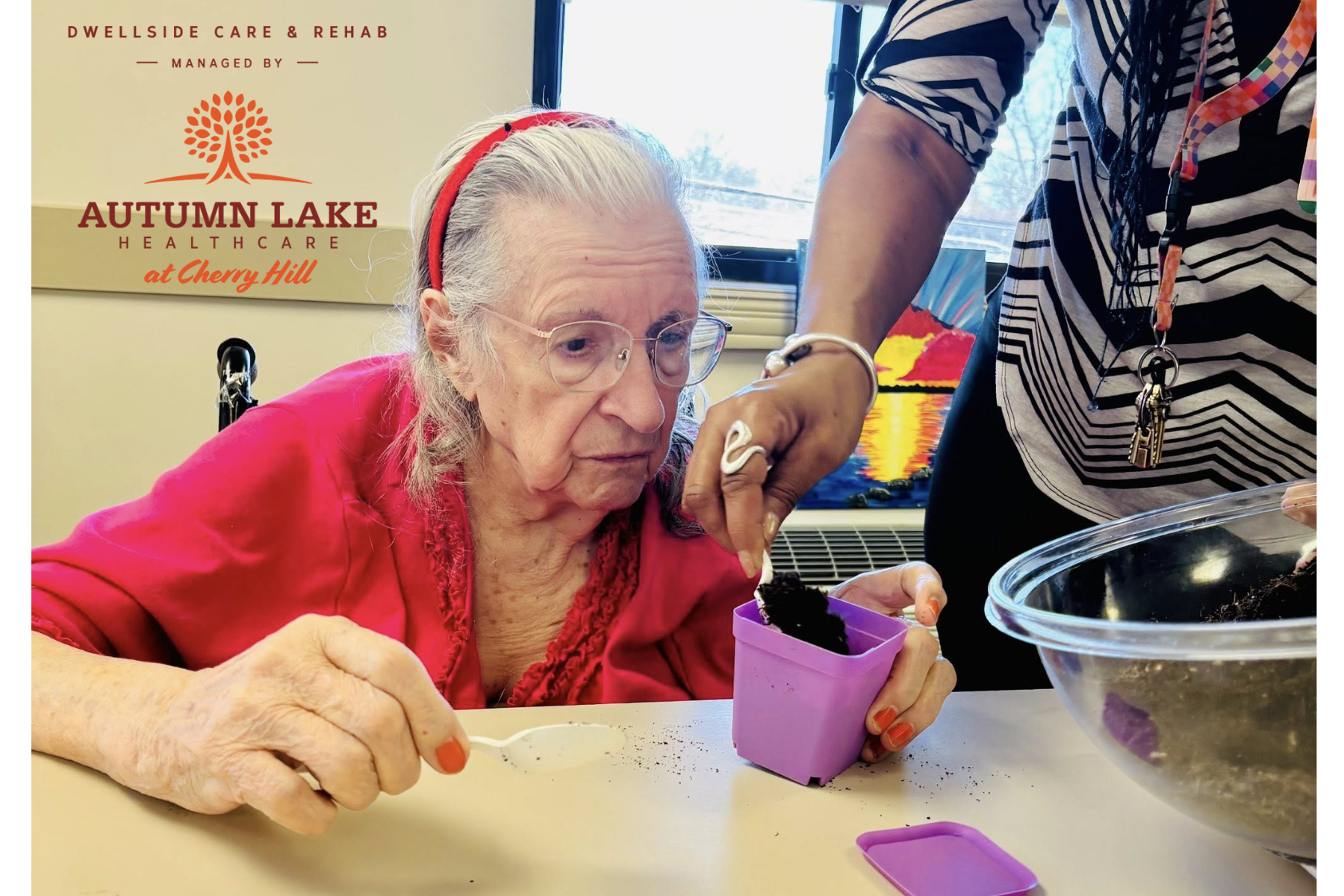 An elderly woman at a rehabilitation center focuses on a therapeutic gardening activity, filling a small purple pot with soil alongside a staff member.