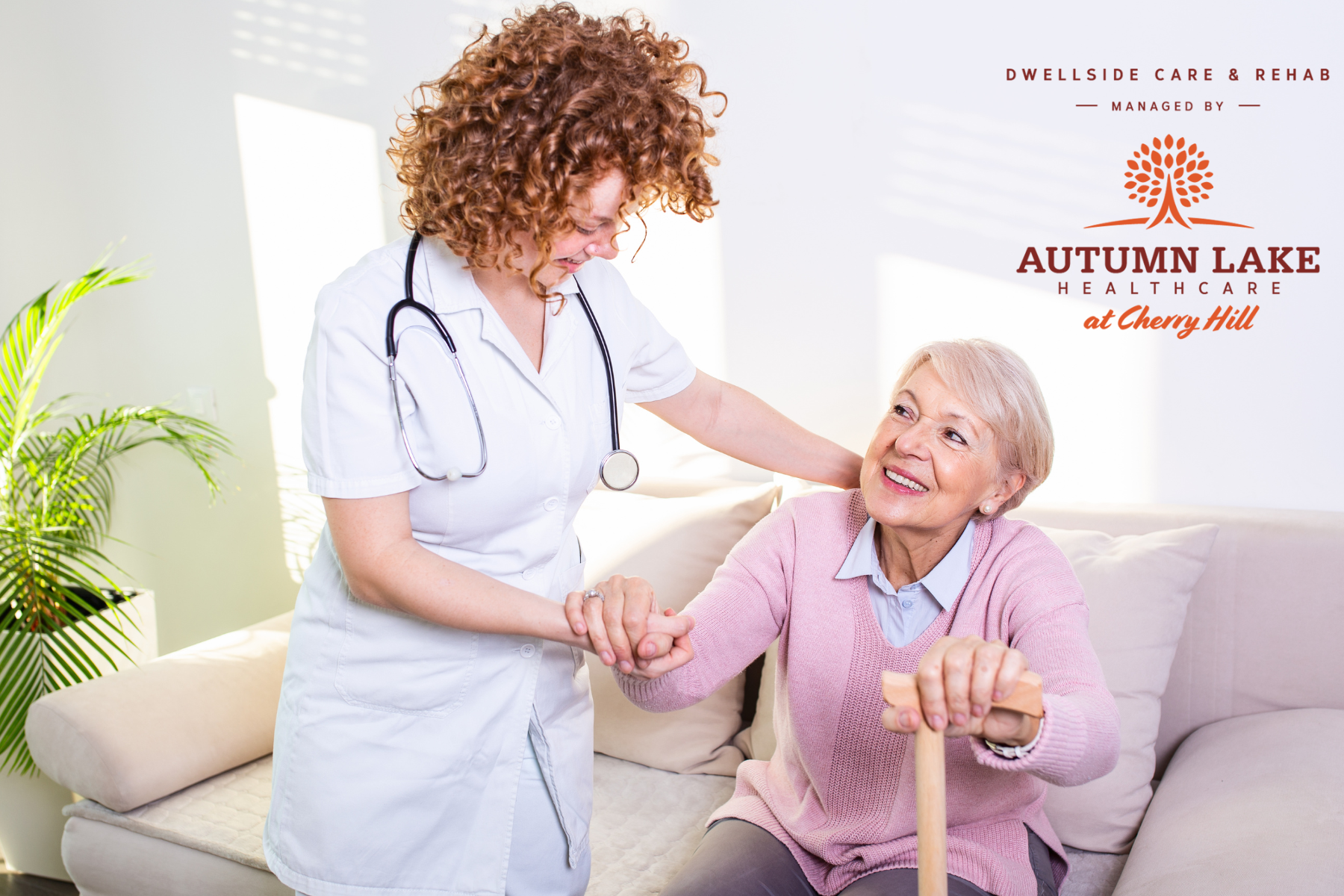 A nurse helps a senior resident stand up from a couch at Autumn Lake Healthcare at Cherry Hill.